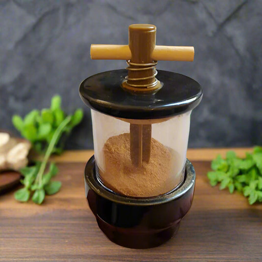 Black herb grinder with ground brown spice in it that you can see through the clear acrylic sides. On a wood table top with green herbs next to it.