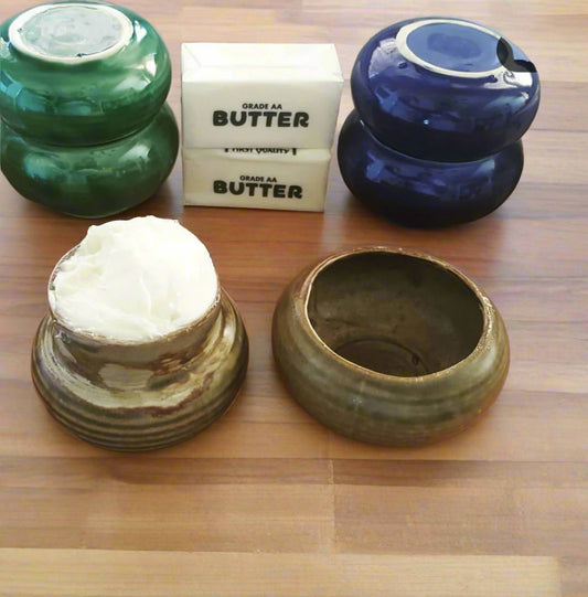 Green, blue and brown butter vases. The brown one has butter in it with the lid next to it. There are 2 sticks of butter on the wood table as well.